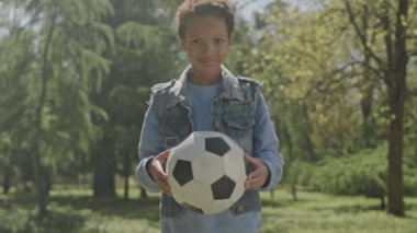 Küçük Afrikalı Amerikalı çocuğun futbol topunu tutarken ve parkta gülümseyerek poz verirken çekilen portre görüntülerini kaldır.