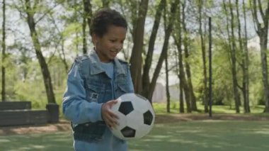 Neşeli Afrikalı Amerikalı çocuk parkta futbol sahasında yürüyor, çimlere top atıyor ve onu tekmelemeye hazırlanıyor.