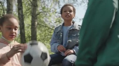 Küçük çocuklar futbol topuyla oynuyorlar, sohbet ediyorlar ve gülüyorlar yaz günü parkta bankta otururken.