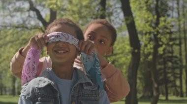 Küçük Afro-Amerikalı kız. Gözleri bağlı gülümseyen arkadaşı. Bir yandan da parkta kör adam taklidi yapmaya hazırlanıyor.