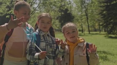Sırt çantaları ve fırıldak oyuncakları olan çok ırklı küçük kızların orta boy portresi. Yaz günü parkta bir arada durup gülümsüyor ve kameraya poz veriyorlar.