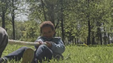 Mutlu küçük çocuk yeşil çimenlerde oturuyor ve arkadaşlarıyla parkta çekiştirirken heyecanla halat çekiyor.