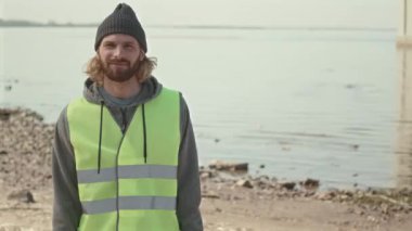 Neon yeşil yelek ve bereli genç gönüllü bir erkeğin orta boy portresi. Sahilde duruyor ve kameraya poz veriyor.