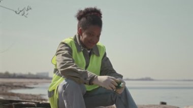 Neon yeşil yelek ve eldivenli, kumsalda oturmuş gülümseyerek kameraya poz veren orta boy siyah kadın çevre gönüllüsü.