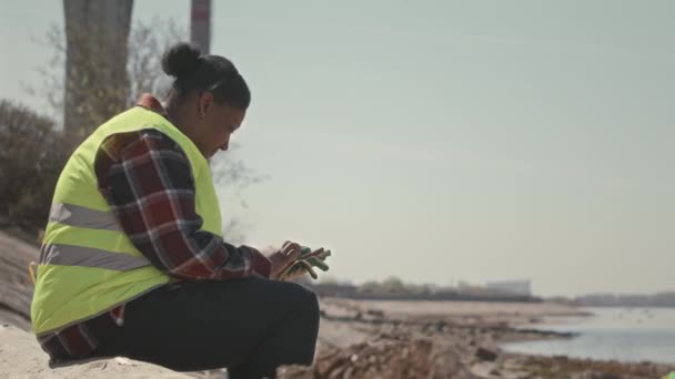 穿着霓虹灯绿色背心的非裔美国女性环保志愿者在海滩上休息并使用智能手机的侧视图 — 图库视频影像