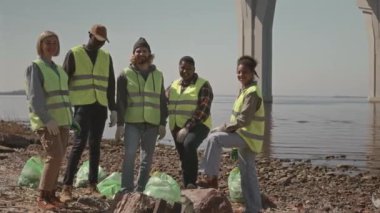 Üniformalı çeşitli çevreci aktivistlerin grup portresi. Kamera önünde birbirlerine poz veriyorlar ve kıyıda çöp torbalarının yanında durup gülümsüyorlar.