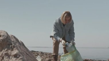 Çöp torbası tutan ve kıyı şeridi temizliği yaparken atık toplayan eldivenli kadın çevreci.