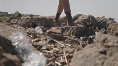 Kayalık sahilde yürüyen ve çöp torbasına plastik atık toplayan çizmeli çevreci aktivistin bacakları.