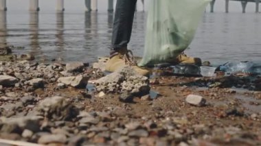 Botlarıyla sahil boyunca yürüyen ve çöp torbasına atık toplayan çevreci aktivistin bacakları.