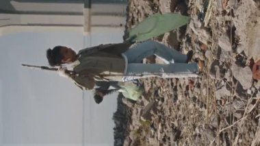 Dikey olarak Afro-Amerikalı Z nesli kızı çöp torbasında çöp toplarken ve sopayla sahilde yürürken çevreci aktivistlerle temizlik yaparken.