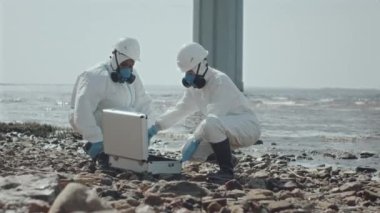 İki ekolojist koruyucu tulumlar ve solunum maskeleri takarak körfez kıyısında çalışırken kirli su örnekleriyle test tüplerini tartışıyor.