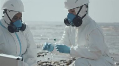Koruyucu tulumlar ve solunum maskeleri takan iki kadın ekolog, test tüplerini kirlenmiş su numuneleriyle inceliyor ve körfez kıyısında çalışırken konuşuyorlardı.