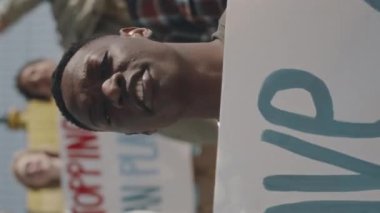 Siyahi bir gencin posterini tuttuğu dikey çekim, kameraya bakıyor ve çevre kirliliğine karşı eylemcilerle protesto ederken slogan atıyor.