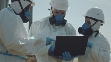 Koruyucu tulumlar, eldivenler, miğferler ve solunum maskeleri takmış profesyonel ekoloji uzmanlarından oluşan bir ekip kirli alanda dizüstü bilgisayar kullanıyor ve konuşuyor.