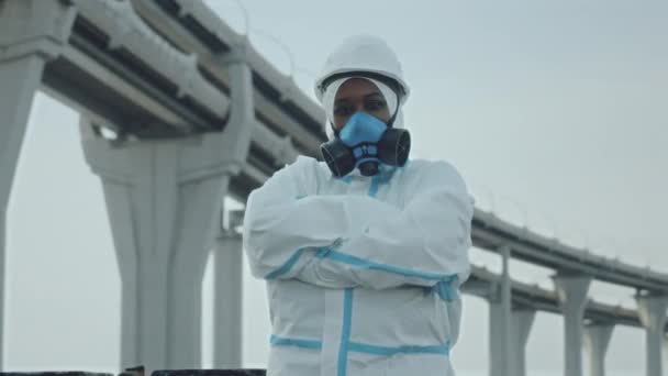 身着防护服 头戴硬帽 头戴呼吸面具 站在室外的非裔美国女生态学家的画像 双手交叉 背景为公路桥梁 摆出相机的姿势 — 图库视频影像