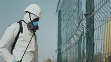 Afrika kökenli, koruyucu örtülü, kalın şapkalı ve solunum maskeli bir çevre bilimci sırt çantası spreyini kullanarak gaz boru hattıyla çitlere kimyasallar yerleştiriyor.