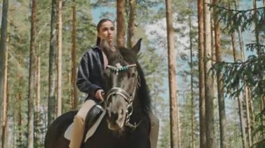 Güneşli bir günde çam ormanında at süren esmer bir kadının düşük açılı çekimi.