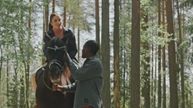 Genç, güzel bir kadın at üstünde oturuyor, yeleyi okşuyor ve ormanda ata binerken Afrikalı Amerikalı erkek arkadaşıyla konuşuyor.