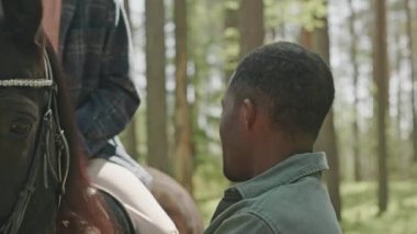 Genç Afrikalı Amerikalı bir adamın at sırtında yelesini sıvazlarken ve ormanda romantik bir randevuda eyerde oturan güzel bir kız arkadaşla konuşurken orta boy yakın çekimi.