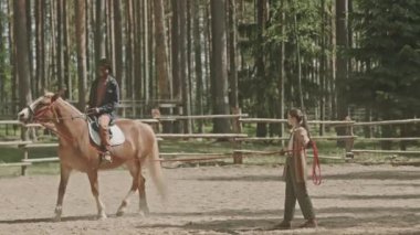 Siyah adamın miğfer takıp ata bindiği geniş açı. Açık hava arenasında, kadın eğitmenin kurşunla tuttuğu ip.