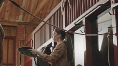 Çiftlikte ahırda çalışırken küçük bir kızın şirin bir atı eyerlerken orta açı çekimi.