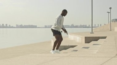 Atletik Afrikalı Amerikalı bir adamın merdivenlerde zıplaması şehrin nehir kıyısında antrenman yaparken çömelmesi.