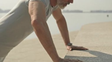 Gri saçlı ve sakallı yaşlı bir adamın sabah şehir nehir kıyısında antrenman yaparken barfiks çekişini çek.