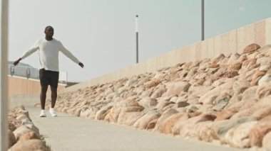 Spor giyimli atletik siyah bir adam kameraya doğru yürüyor, kollarını sallıyor ve antrenmandan önce ısınma egzersizleri yapıyor.