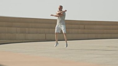 Spor kıyafetlerinde enerjik kıdemli bir adam atlıyor ve dışarıda sabah antrenmanına hazırlanırken omuzlar ve boyun için ısınma egzersizleri yapıyor.