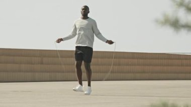 Açık havada kardiyo antrenmanı yaparken şehirde spor kıyafetleriyle ip atlayan Afro-Amerikan bir adamın geniş açı çekimi.