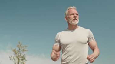 Düşük açılı yavaş çekim. Sporcu kıdemli sporcu güneşli bir yaz sabahı bulutsuz gökyüzüyle dışarıda koşuyor.