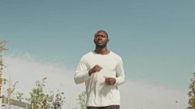 Düşük açılı ağır çekim. Atletik siyah adam sabah antrenman yaparken dışarıda koşuyor. Bulutsuz gökyüzü.