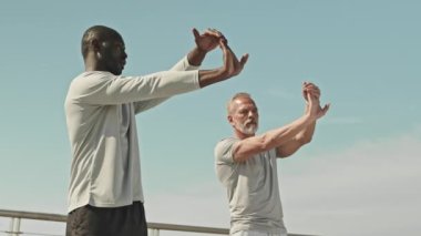 Açık hava spor antrenmanından önce ısınırken, yaşlı, kır saçlı bir adama bilek esnetme egzersizi yapan siyah antrenörün düşük açılı çekimi.