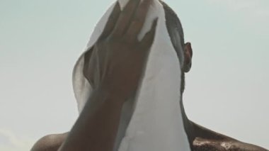 Üstsüz Afrikalı Amerikalı sporcunun güneşli bir günde açık havada yapılan yüksek yoğunluklu egzersizden sonra tenindeki teri havluyla silerkenki düşük açılı görüntüsü.