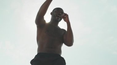Gömleksiz, kaslı siyah bir adamın açık havada güneşli bir günde spor yaparken gölgede boks yaparken, gün ışığı kameraya yansıyor.