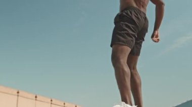 Alçak açılı, üstsüz, Afrika kökenli Amerikalı bir adamın yaz günü şehirde antrenman yaparken bank atlayışı yaptığı bir sahne.