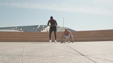 Üstsüz, yaşlı bir adamın şehirde spor yaparken genç siyahi bir antrenörle çadır kurması.