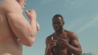 Gövdesi kaslı siyah bir adamın açık havada boks dersi verirken üst düzey bir adama nasıl yumruk atacağını açıklarken alçak açılı çekimi.