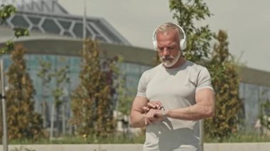 Kablosuz kulaklıklı orta yaşlı bir adamın spor takip uygulamasını akıllı kasete yerleştirirken orta boy fotoğrafı. Sonra da şehirde koşmaya başlarsın.