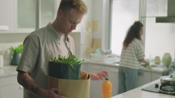 Молодий Чоловік Приносить Пакет Продовольства Знімає Кухонний Стіл Спілкується Своєю — стокове відео