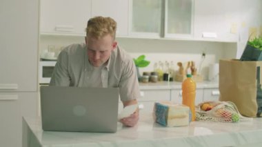 Genç bir adamın mutfak masasına eğildiği orta boy bir fotoğraf, alışveriş fişi okurken ve evde kişisel bütçesini yönetirken dizüstü bilgisayar kullanırken.