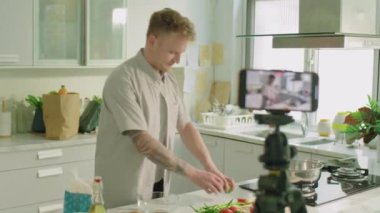 Genç erkek yemek blogcusu mutfakta dikilirken, kamerada sebze gösterip yemek tarifini anlatırken, akıllı telefonuyla tripodla video tarifi çekerken orta ölçekli bir fotoğraf.