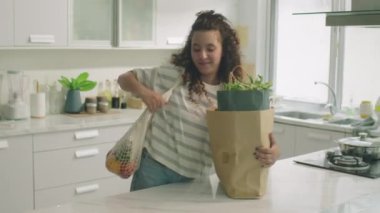 Genç kıvırcık saçlı kız alışveriş çantalarını evdeki mutfak masasına getiriyor ve marketten alışveriş torbalarını boşaltıyor.