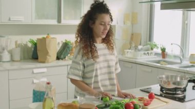 Orta boy kıvırcık saçlı bir kız mutfakta dikilirken, sebze ve bıçak gösterirken ve yemek videosu çekerken yemek tarifini anlatırken.