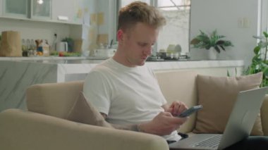 Genç adamın evdeki kanepede oturmuş online alışveriş yaparken ya da faturaları öderken bilgisayarını ve akıllı telefonunu kullanırken orta boy bir fotoğrafı.
