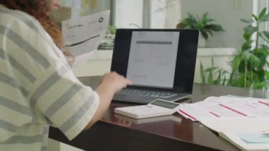 Evdeki faturaları öderken dizüstü bilgisayar ve hesap makinesi kullanan genç bir kadının omuz üstü fotoğrafı.