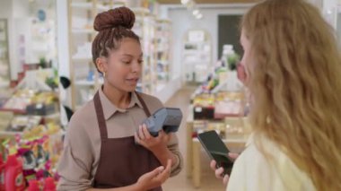 Genç kozmetik mağazası çalışanının ürünleri satın almak için akıllı telefonu kullanan kadın müşteriye ödeme terminali verdiği orta boy bir fotoğraf.