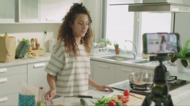 Kızın mutfakta durup, yemek malzemelerini gösterirken ve kamerada konuşurken tripodla akıllı telefon ile video bloğu çekerken orta ölçekli görüntüsü.