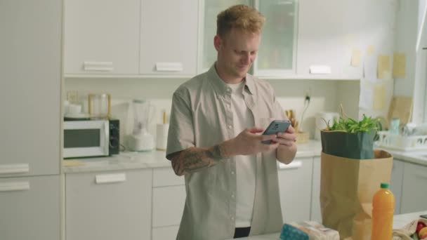 Genç Adam Elinde Alışveriş Poşetleriyle Mutfakta Duruyor Akıllı Telefon Kullanıyor — Stok video