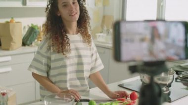 Genç bayan yemek blogcusu mutfakta dikilirken, kamerada sebze gösterirken ve yemek tarifini anlatırken, akıllı telefonuyla video blogu çekerken orta boy bir fotoğraf.